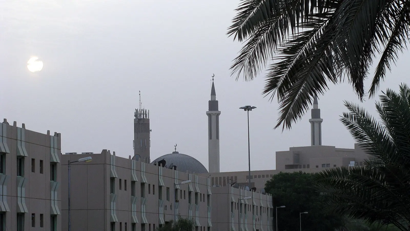 منظر مسائي لحيّ سكني في الرياض بمآذن مسجد وسيارات مصفوفة تحت نخيل، والشمس تغرب خلف المباني.